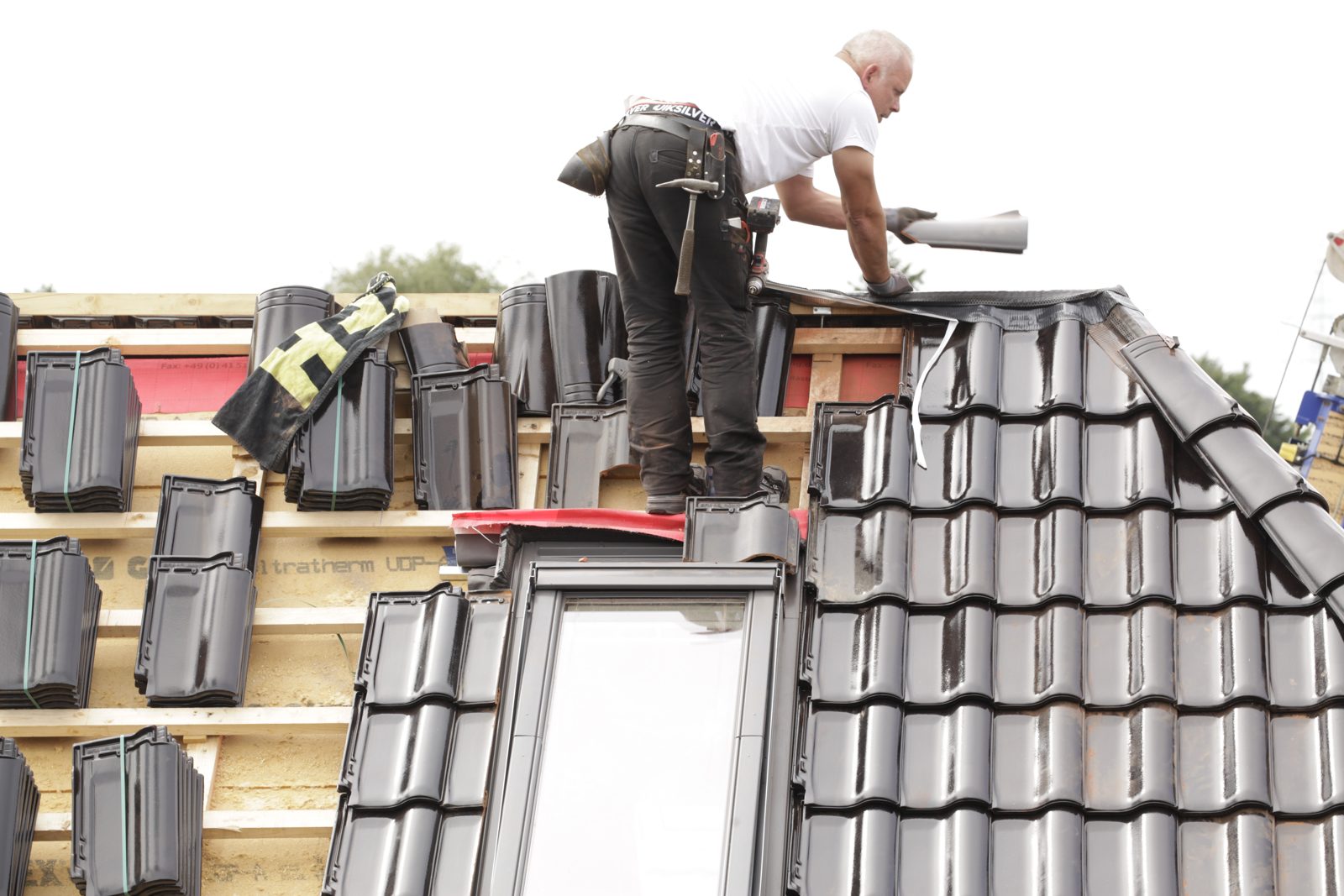 A roofer in a white shirt installing dark roof tiles around a skylight on a pitched roof