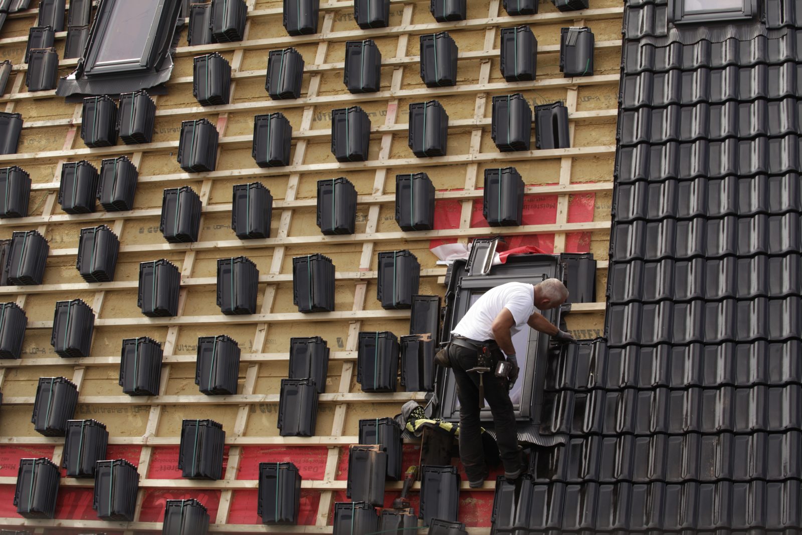 A roofer installing dark roof tiles next to a skylight on a partly finished roof