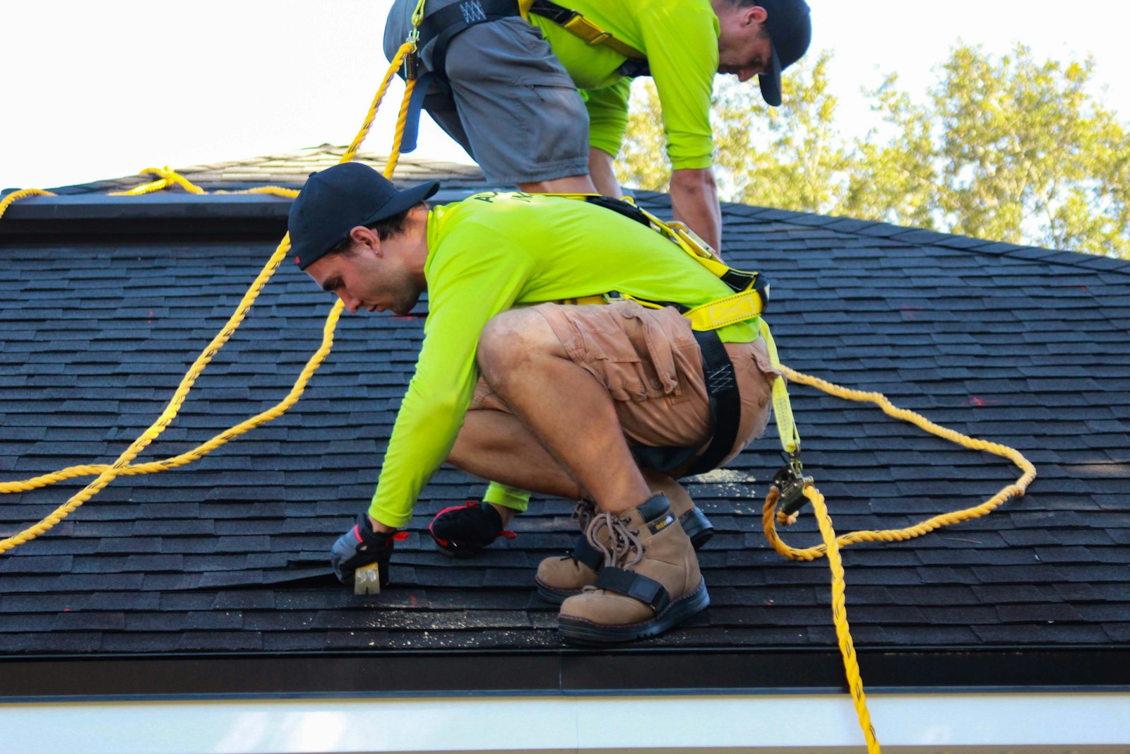 Two roofers in safety harnesses repairing shingles on a house roof